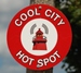 Round sign. The outside circle has a red background and in white letters it reads: "Cool City Hot Spot." In the middle of the circle against a white background is a picture of a red tower with a grey signal coming from it.